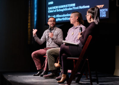 Two men and a woman are seated on stage, holding microphones and talking