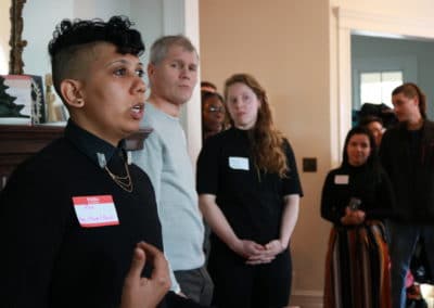 A non-binary person dressed in black talks in front of a fireplace while others listen