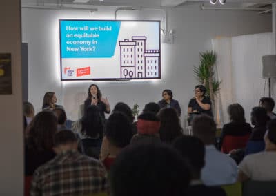 view from the crowd of four people on stage with a powerpoint that reads "How will we build an equitable economy in New York" above them