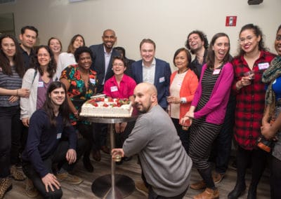 A large group of diverse people from all backgrounds smiling huddles around a cake