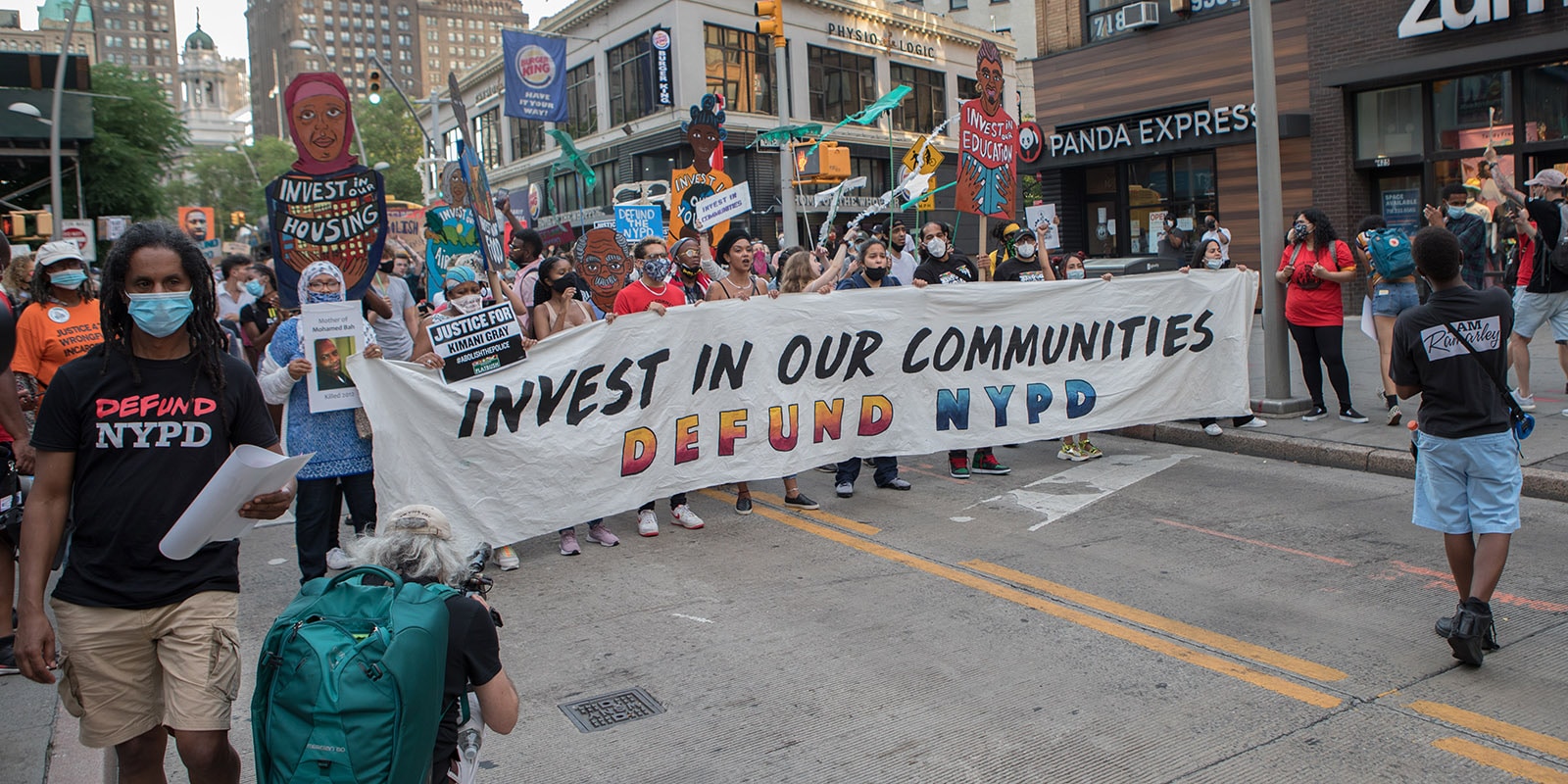 People marching in the street with masks and a banner that say, "Invest in our Communities, Defund NYPD"