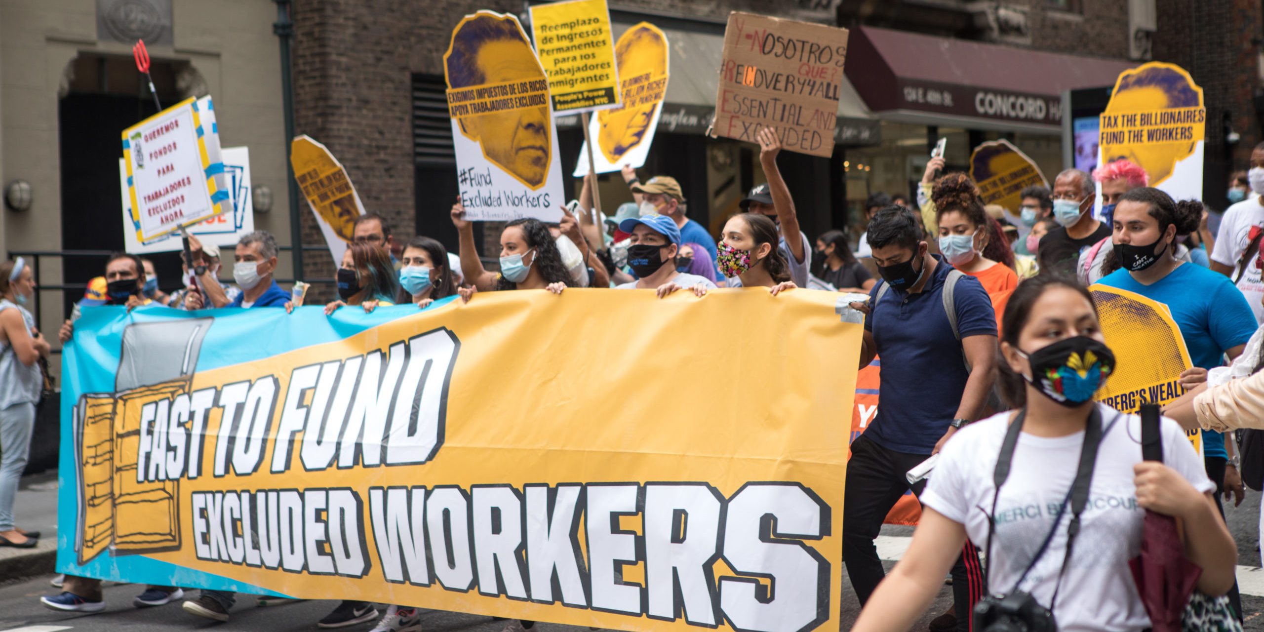Outdoor protesters with masks and a banner that reads "Fast to Fund Excluded Workers"